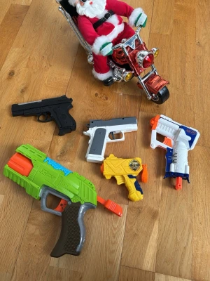 Leksakspistoler och tomte på motorcykel - Säljer ett set med fem olika leksakspistoler i plast i färgerna svart, grå, vit, grön, orange, gul och blå. Medföljer även en tomtefigur sittandes på en röd motorcykel. Perfekt för lek och fantasi. Tomten kostar 50kr leksakspistolerna kostar 30kr/st
