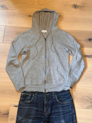 Grå zip hoodie från Davida - Mjuk och stilren grå hoodie från Davida med dragkedja framtill och två fickor. Tillverkad i ett skönt stickat material, troligen ull eller kashmir. Klassisk huva med dragsko och ribbade muddar vid ärmslut och nederkant. Perfekt för en avslappnad look.