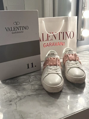Valentino Rockstud Untitled sneakers - Snygga vita sneakers från Valentino Garavani med ljusrosa band och ikoniska nitar över ovandelen. Skorna har en klassisk låg siluett, vita snören och detaljerade kanter. Tillverkade i läder för en lyxig känsla och stilren look.                               Jag köpte skorna på vinted men säljer eftersom de tyvärr va för små! Men de är i mycket bra skick💗💗