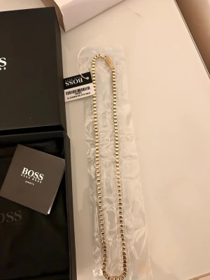 Guldigt halsband från Hugo Boss - Säljer ett stilrent halsband från Hugo Boss i guldtonad metall. Halsbandet har en kedja med fyrkantiga länkar som ger en modern och elegant look. Perfekt för dig som vill ha ett exklusivt smycke med snygg design. Levereras i originalask med Boss-logga. Den är lång 