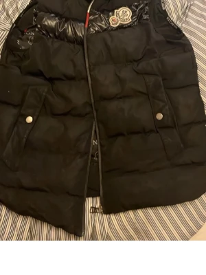 Svart dunväst från Moncler - Säljer en svart dunväst från Moncler med quiltad design och glansiga detaljer upptill. Västen har ett klassiskt snitt och är tillverkad i polyester med dunfyllning. Insidan har Moncler-loggor tryckta över hela fodret för en lyxig känsla. Perfekt för kalla dagar.