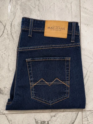 Mörkblå raka jeans från MAC Jeans - Snygga mörkblå jeans från MAC Jeans med raka ben och klassisk femficksmodell. Jeansen har kontrastsömmar och en cool detalj på bakfickan. Tillverkade i ett slitstarkt denimtyg som passar perfekt till din vardagsstil.33/32