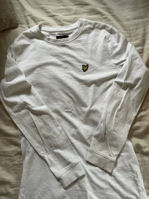 Vit långärmad t-shirt från Lyle & Scott - Vit långärmad t-shirt från Lyle & Scott med klassisk rund halsringning och den ikoniska gula örnloggan broderad på bröstet. Tillverkad i mjuk bomull som känns skön mot huden. Perfekt för en clean och stilren look.
