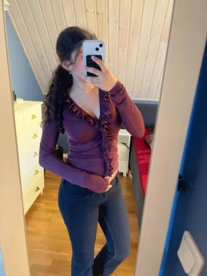 Vinröd blus med volangdetaljer - Säljer en vinröd blus med långa ärmar och djup V-ringning. Blusen har snygga volangdetaljer längs ringningen och framtill, vilket ger en trendig och feminin look. Materialet är stretchigt och sitter figurnära för en smickrande passform.