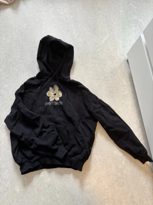 Svart hoodie med blomma och text - Svart hoodie med huva och magficka. På bröstet finns en stor gul och vit blomma samt texten 'SWEET SKTBS'. Hoodien har ribbade muddar och är i mjukt material, perfekt för en chill stil.
