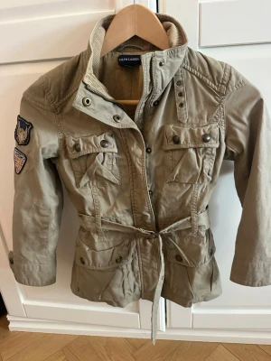 Vår/höstjacka  från Ralph Lauren - Stilren beige jacka från Ralph Lauren med coola patchar på ärmen, fyra fickor framtill och knytskärp i midjan. Jackan har hög krage, tryckknappar och dragkedja. Perfekt för en trendig streetlook.Finns på Östermalm . Nypris var 3600 på nk 