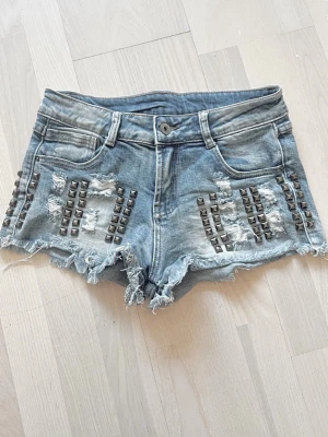 Blå jeansshorts med nitar och slitningar - Snygga blå jeansshorts med råa kanter, slitningar och coola nitar framtill. Klassisk femficksmodell med låg midja och fransig nederkant. Perfekta för en edgy streetstil och sommarens alla äventyr.