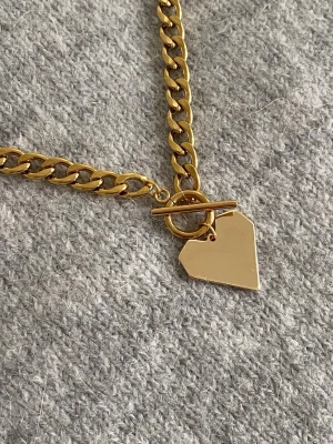 Guldigt halsband med hjärthänge - Snyggt halsband i guldfärgad rostfritt. Kedjan är chunky och har ett geometriskt hjärthänge samt ett dekorativt T-lås framtill. Perfekt accessoar för att lyfta din outfit med en trendig touch.