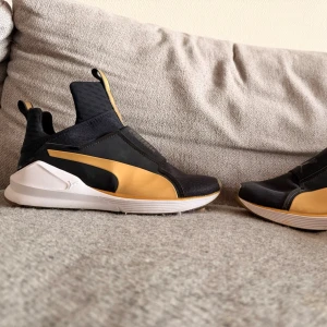 Svarta och guldiga sneakers från Puma - Snygga sneakers från Puma i svart med guldiga detaljer och vit sula. Skorna har en modern slip-on design utan snörning, högre skaft och elastiska partier för enkel påtagning. Materialet är en mix av textil och syntet. Perfekta för dig som gillar sportig och trendig stil.