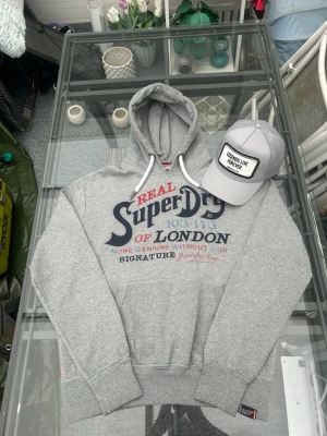 Superdry Hoodie Grå Large – Clean Summer Streetwear Y2K - äljer en snygg grå Superdry hoodie i storlek L. Hoodien är i mycket bra skick utan defekter och passar perfekt till sommar kvällar, streetwear outfits och casual fits. Mjuk och bekväm med fleecefodrad insida samt stor Superdry logga framtill som ger en clean vintage/Y2K look. Tillverkad i bomullsmix med hög komfort och bra passform. Perfekt hoodie för everyday wear, skate, relaxed fit och layered outfits. 🔥  • Märke: Superdry • Storlek: L • Färg: Grå • Skick: Mycket bra skick, inga defekter 