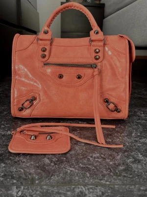 Korallrosa handväska  - Fin orange/ rosa handväska,  dragkedja framtill och flätat handtag. Väskan är i glansigt läder och har en tillhörande liten accessoar. Perfekt för dig som vill sticka ut med en färgstark och trendig väska.