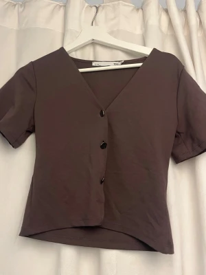 Brun v-ringad topp med knappar - Kortärmad brun topp från lager 157med v-ringning och tre svarta knappar framtill. Modellen är croppad och har en enkel, stilren design som passar perfekt till jeans eller kjol.