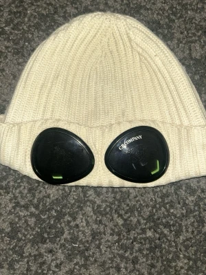 Vit cp company mössa - Säljer en vit ribbstickad mössa från C.P. Company med de ikoniska svarta goggles framtill. Mössan är tillverkad i mjukt stickat material och har en uppvikt kant. Perfekt för dig som vill sticka ut med en streetwear-inspirerad look.