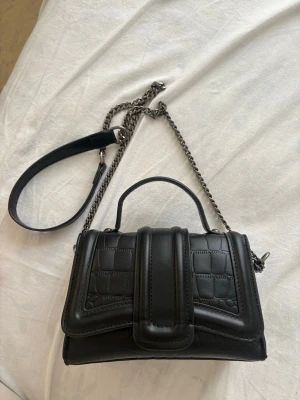 Svart croco handväska med kedja Zara - Snygg svart handväska i croco-mönstrat konstläder med silverfärgad kedjerem och ett stilrent handtag. Väskan har en unik frontdetalj och passar perfekt för dig som gillar trendiga accessoarer. Rymmer det viktigaste och ger en cool vibe till din outfit.