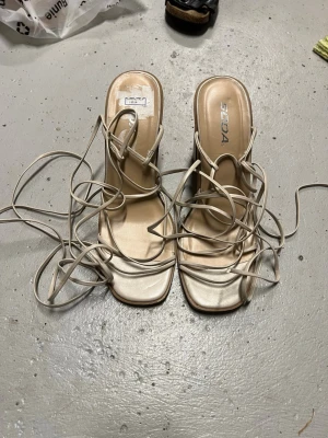 Beige snörade sandaler - Stilrena beige sandaler med fyrkantig tå och tunna remmar som snöras runt vristen. Klackarna är medelhöga och ger en trendig look. Tillverkade i syntetmaterial med en elegant finish. Perfekta för dig som gillar minimalistisk och modern stil. Nya!