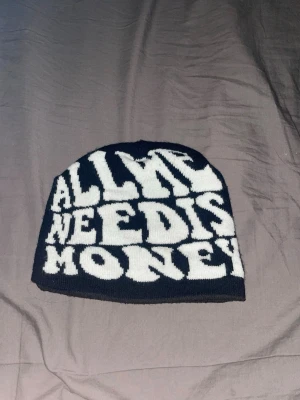 Svartvit mössa med textmotiv - Cool svart mössa med vit text som säger 'ALL WE NEED IS MONEY' i stora bokstäver. Mössan är stickad och har en avslappnad passform, perfekt för att ge din outfit en edgy touch.