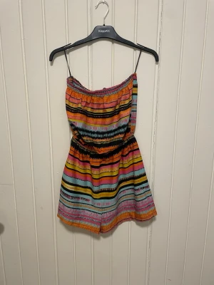 Färgglad randig jumpsuit med tunna axelband - Säljer en superfin jumpsuit med färgglada ränder i orange, rosa, gult, svart och turkost. Modellen har tunna axelband, resår i midjan och är kort i benen. Perfekt för dig som gillar färg och mönster! Materialet känns lätt och luftigt.