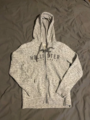  hoodie från Hollister - Snygg hoodie från Hollister med dragkedja framtill och justerbar huva. Tröjan har långa ärmar, fickor på sidorna och tryckt logga över bröstet. Perfekt för en avslappnad och casual stil.