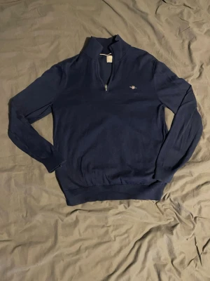 Mörkblå halvzip-tröja från GANT - Mörkblå långärmad tröja från GANT med halv dragkedja och liten broderad logga på bröstet. Tröjan har hög krage och ribbade muddar vid ärmslut och nederkant. Perfekt för en stilren och avslappnad look.