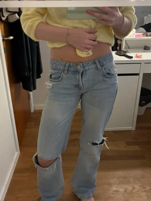 Ljusblå slitna jeans med hål - Säljer ett par ljusblå jeans med raka ben och slitna detaljer, inklusive stora hål vid knäna. Jeansen har klassisk femficksdesign och låg midja. Perfekta för en avslappnad och trendig look.