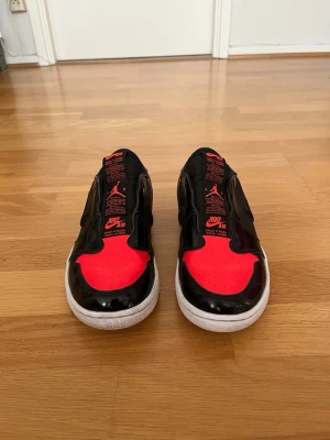 Nike Air Jordan 1 Low Slip - storlek 39 - Säljer ett par snygga och stilrena Nike Air Jordan 1 Low Slip i svart och rött. Skorna är i bra använt skick med normalt slitage och fungerar perfekt både till vardag och streetwear. Storlek 39 och väldigt bekväma på foten. Den klassiska Jordan-designen med röda detaljer gör att de sticker ut riktigt fint.