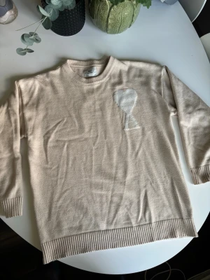 Beige stickad tröja Ami Paris - Snygg beige stickad tröja från Ami Paris med det ikoniska vita hjärta-A-motivet på bröstet. Tröjan har rund hals, ribbade muddar och är tillverkad i mjukt material som känns skönt mot huden. Perfekt för dig som gillar stilrena och trendiga plagg.
