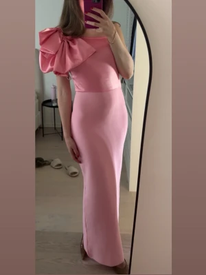 Rosa långklänning med rosett - Supersnygg rosa långklänning i satinliknande material med en stor rosett på ena axeln. Klänningen är oneshoulder. Från Bella Barnett. Använd 2 gånger! 