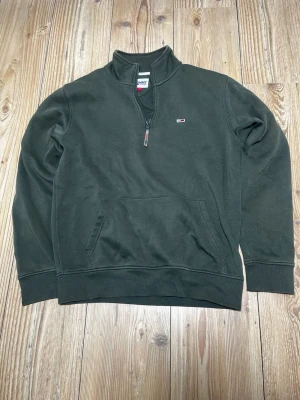 Mörkgrön halfzip hoodie Tommy Hilfiger - Mörkgrön hoodie från Tommy Hilfiger med halv dragkedja och klassisk logga på bröstet. Tröjan har känguruficka framtill, ribbade muddar och hög krage. Tillverkad i mjuk bomullsblandning som är skön att bära.