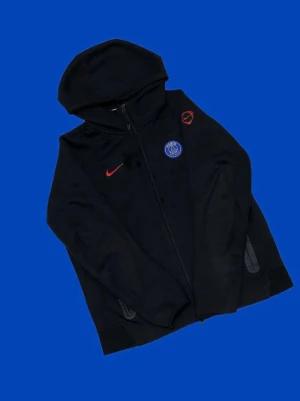 Svart PSG Nike tech från Nike - Svart hoodie med dragkedja från Nike och PSG-logga på bröstet. Röd Nike swoosh och PSG-märke framtill. Utrustad med huva och fickor på sidorna. Perfekt för dig som gillar sportig stil och vill representera Paris Saint-Germain.