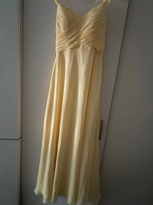 Ljusgul långklänning med öppen rygg - Superfin ljusgul långklänning med smala axelband och vackert draperad överdel. Klänningen har öppen rygg med snörning och knyt, vilket ger en romantisk och somrig känsla. Perfekt för speciella tillfällen eller sommarens fester.