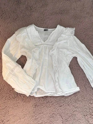Vit volangblus från Gina Tricot - Supersöt vit blus från Gina Tricot med volangdetaljer och spets längs kanterna. Blusen har v-ringning, långa ärmar och romantisk känsla. Perfekt att styla med jeans eller kjol för en trendig look. Passar även xxs-s och säljs inte längre i butik så passa på att köpa för ett sjukt billigt pris‼️(köpte för 299kr). Använts 1 gång.