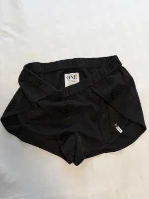 Svarta shorts från ONE - Svarta shorts från ONE med elastisk midja och diskret ormmönster. Modellen är sportig med omlott-detaljer i sidorna och tillverkad i ett lätt syntetmaterial som andas. Perfekta för dig som gillar att röra på dig och vill ha en cool look 🤍
