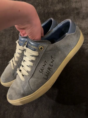 Saint Laurent grå mocka sneakers - Gråa sneakers i mocka från Saint Laurent med handskriven logga på sidan, vita snören och gulaktig sula. Hälpartiet är i blått läder och skon har en klassisk låg profil. Perfekt för dig som gillar stilrena och exklusiva sneakers.