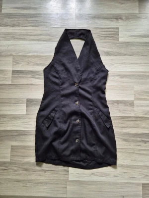 Svart kort klänning med knappar - Snygg svart kort klänning med djup v-ringning och knäppning framtill. Klänningen är ärmlös och har två fickor framtill. Perfekt för dig som gillar en stilren och modern look.