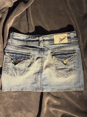 Ljusblå jeanskjol från Guess - Ljusblå kort jeanskjol från Guess( storlek 26)  med klassiska fickor fram och bak, bälteshällor och snygga knappdetaljer. Kjolen har en tydlig logopatch bak och är gjord i bomull. Perfekt för dig som gillar denim och vill ha en trendig look.  