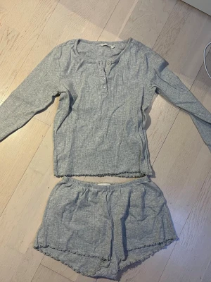pyjamas - Mysig grå pyjamas i två delar med långärmad tröja och shorts. Tröjan har knappar framtill och båda delarna har diskreta volangdetaljer vid kanterna. Ribbat tyg för extra komfort, perfekt för hemmakvällar.