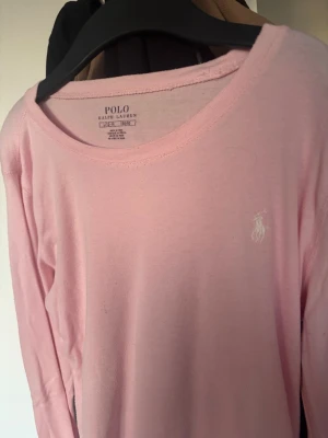 Ljusrosa långärmad topp Polo Ralph Lauren - Supersöt ljusrosa långärmad topp från Polo Ralph Lauren med rund halsringning och diskret broderad logga i vitt på bröstet. Tillverkad i mjukt bomullsmaterial som känns skönt mot huden. Perfekt att styla med jeans eller kjol.