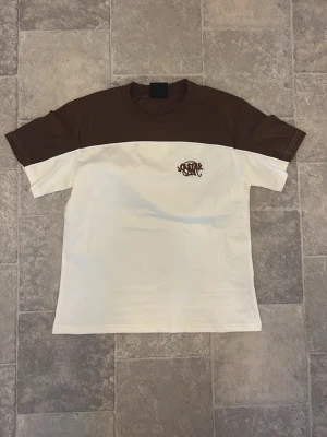 Brun och vit t-shirt från syna world  - Snygg t-shirt från syna world i tvåfärgad design med brun överdel och ärmar samt vit nederdel. T-shirten har rund halsringning och korta ärmar. Liten logga på bröstet. Perfekt för en chill och stilren look. Ej använd 