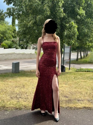 Vinröd glittrig långklänning med slits - Vinröd långklänning med glittrigt tyg, smala axelband och snörning i ryggen. Klänningen har en hög slits framtill och figurnära passform. Perfekt för bal eller festliga tillfällen. Använd 1 gång. 