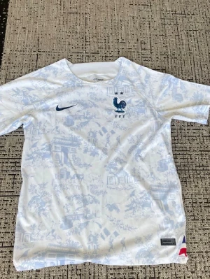 Frankrike Nike fotbollströja vit/blå - Säljer en officiell Frankrike fotbollströja från Nike i vitt med ljusblått mönster. Tröjan har korta ärmar, rund hals och FFF-logga med tupp på bröstet samt Nike Swoosh. Tillverkad i lätt och ventilerande material, perfekt för träning eller match.