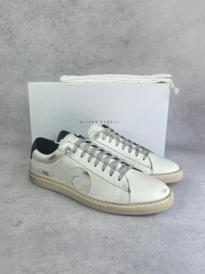 Vita Oliver Cabell sneakers - Stilrena vita sneakers från Oliver Cabell med grå snörning och svart hälparti. Skorna har en klassisk låg siluett och är tillverkade i läder med diskret cirkeldetalj på sidan. Perfekta för en clean och modern look.