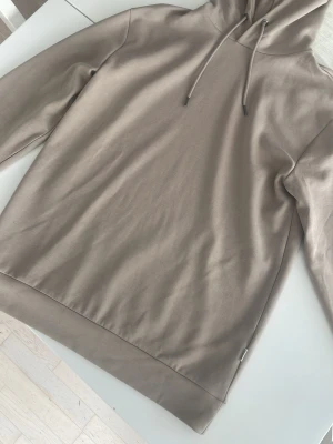 Beige hoodie från Selected Homme - Snygg beige hoodie från Selected Homme i storlek L. Hoodien har en klassisk design med dragsko i huvan och ribbade muddar. Tillverkad i mjukt material som känns skönt mot huden. Perfekt för en avslappnad och stilren look.