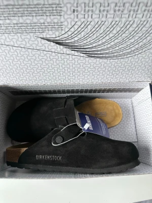 Svarta Birkenstock Boston tofflor - Säljer ett par svarta Birkenstock Boston tofflor i mocka med klassisk korksula och justerbart spänne. Tofflorna har en mjuk innersula i ljust material och är perfekta för en chill och stilren look. Märket är tryckt på sidan och de kommer i originalkartong.