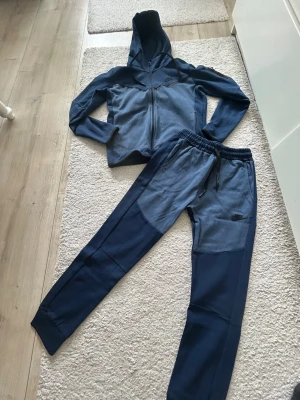 Blått Nike loungewear set - Mysigt tvådelat loungewear set från Nike i två nyanser av blått. Setet består av en hoodie med dragkedja och justerbar huva samt matchande byxor med resår och dragsko i midjan. Båda delarna har diskret Nike-logga och är tillverkade i mjukt material.
