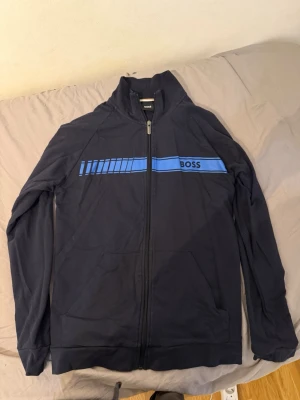 Hugo Boss hoodie - Snygg mörkblå hoodie från BOSS med dragkedja och blått tryck över bröstet. Hoodien har två fickor framtill och en stilren design utan synliga loggor på ryggen. Tillverkad i mjukt material som känns skönt mot huden. Perfekt för en avslappnad och trendig look.