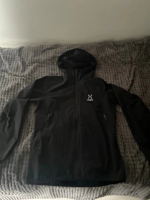 Svart vindjacka från Haglöfs med huva - Svart vindjacka från Haglöfs med hel dragkedja, två sidofickor med dragkedja och justerbar huva. Jackan har minimalistisk design och är tillverkad i ett lätt och smidigt material som passar perfekt för blåsiga dagar. Det är size M men det passar S