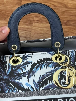 Dior handväska med blått mönster - Snygg handväska från Dior med blått och vitt tropiskt mönster, mörkblå handtag och guldfärgade metalldetaljer. Väskan har klassiska Dior-hängen och är tillverkad i canvas med exklusiv känsla. Perfekt för dig som vill sticka ut med en ikonisk accessoar. A kopie väska 