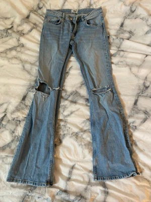 Ljusblå bootcut jeans med slitningar - Säljer ett par ljusblå bootcut jeans från lager157 med slitna detaljer och hål på båda knäna. Jeansen har klassisk femficksdesign, normal midja och dragkedjegylf. Perfekta för en avslappnad och trendig look.