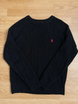 Svart stickad tröja från Ralph Lauren - Svart stickad tröja från Ralph Lauren med klassiskt kabelstickat mönster och röd broderad logga på bröstet. Tröjan har rund halsringning och långa ärmar. Perfekt för dig som gillar stilrena och tidlösa plagg.