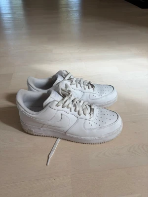 Nike Air Force 1 vita sneakers - Klassiska vita Nike Air Force 1 sneakers i lågt utförande. Skorna är tillverkade i läder med perforerad tåbox för ventilation, vit sula och matchande vita skosnören. Swoosh-logga på sidan och diskret branding på plösen och hälen.